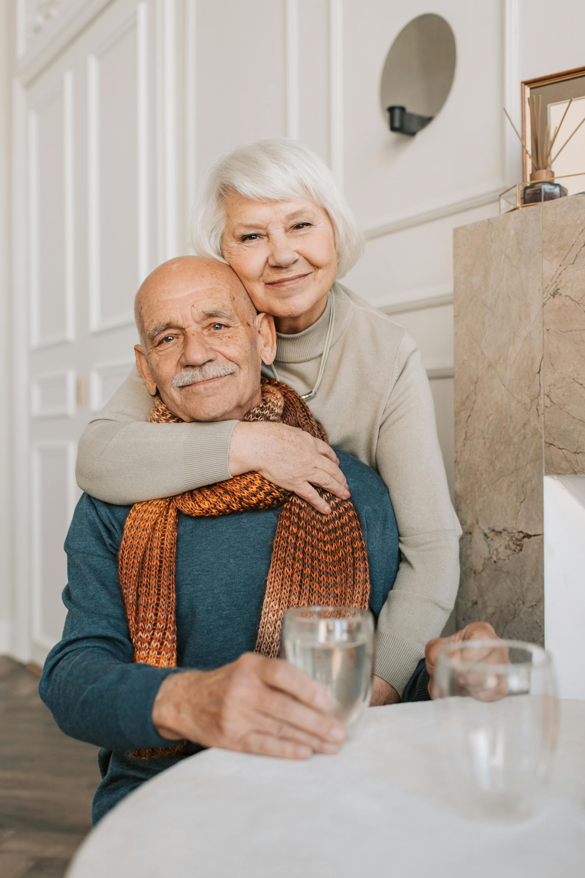 An older couple at home, the woman warmly leaning over the man's shoulder, evoking the hospitality of well-run senior living